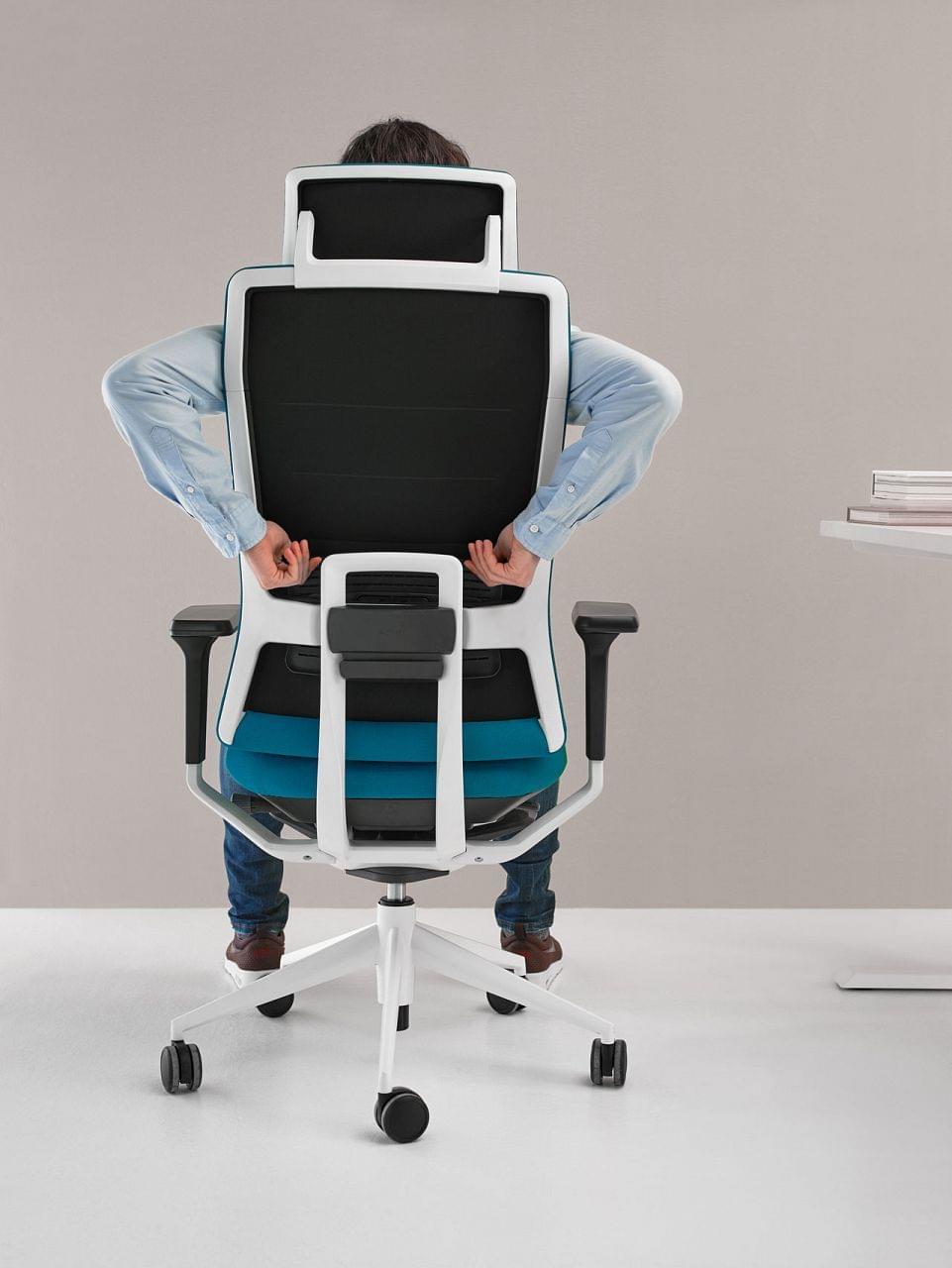 Polohovatelná černo-šedá kancelářská židle s prvky tyrkysové barvy. Obrázek je zaměřen na zaměstnance sedícího na ergonomické židli, který si nastavuje opěrku zad. Kancelářské židli nechybí ani otáčecí kolečka pro změnu polohy na pracovišti. 
