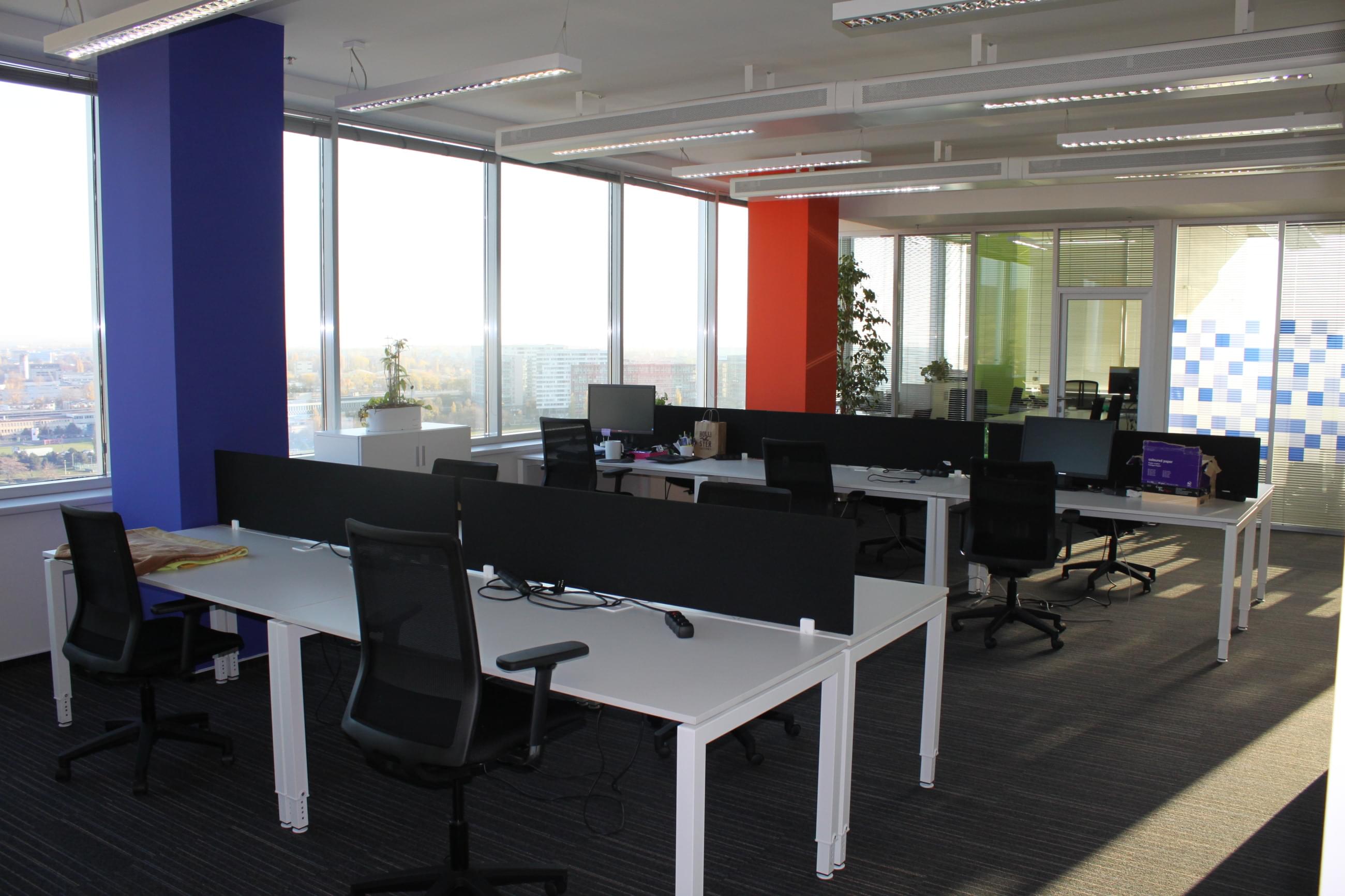 Pohled do openspace kancelacáří společnosti Concentrix s provními stoly v půli dělenými černou přepážkou. Prostoru dominují velké sloupy u stěny s okny, které jsou různě barevné.
