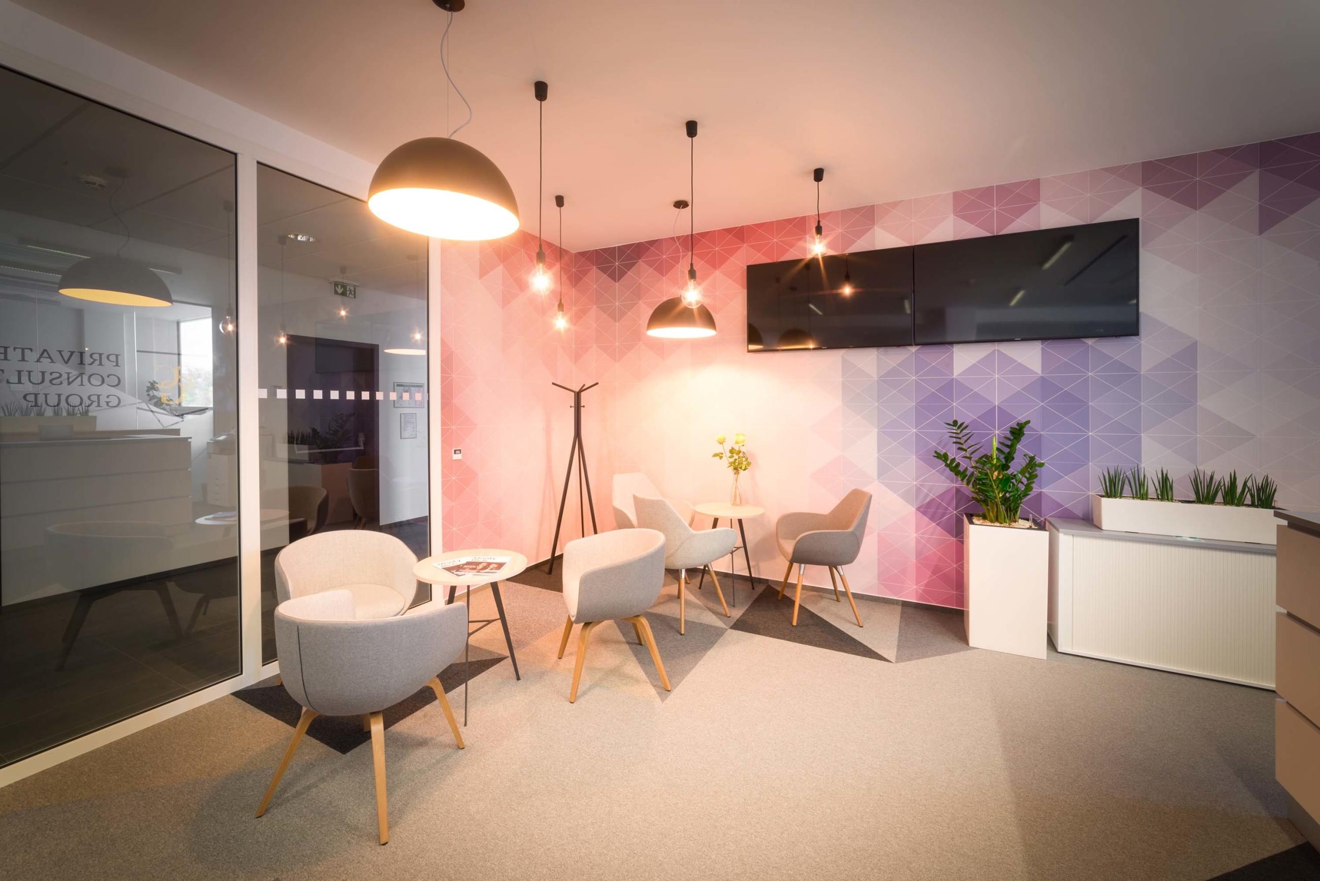 Šedá křesílka s malými stolečky jako místo k setkávání s LCD obrazovkami a zajímavě řešeným osvětlením a barevnou tapetou v pozadí kontrastní k jinak šedému interiéru.Šedá křesílka s malými stolečky  doplňuje zajímavě řešené osvětlení a barevná tapeta, která je kontrastní k jinak šedému interiéru.