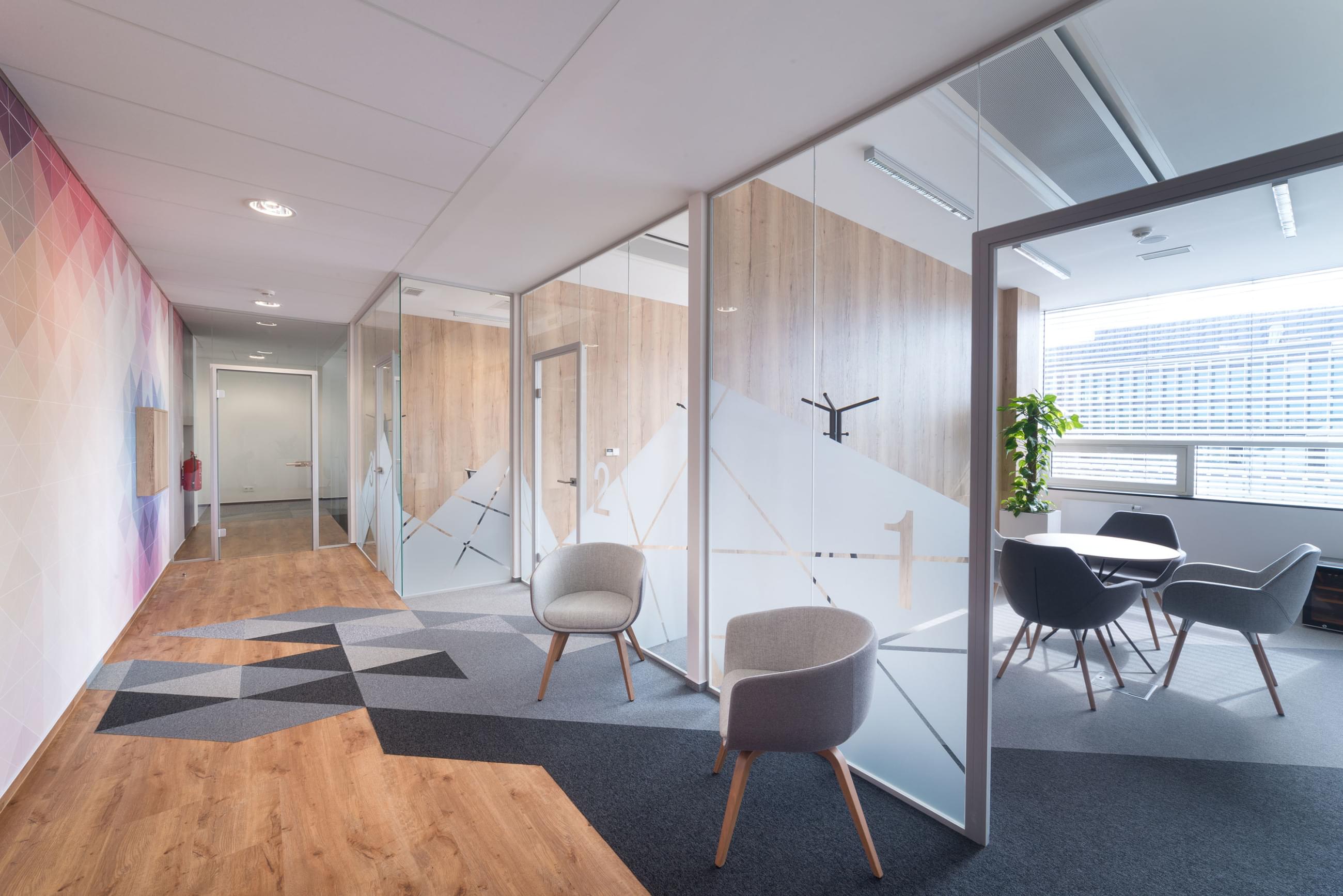 Pohled do chodby zachycující podlahu řešenou kombinací šedých kobercových dlaždic  s vinylem, skleněné příčky s polepy a dvě stoličky vše sladěné do šedých tónů.