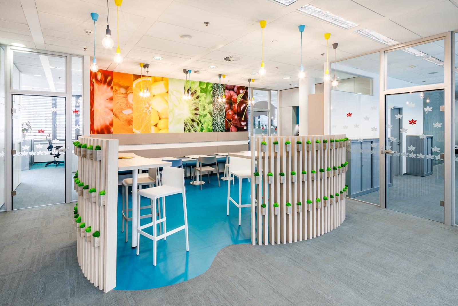 Barevná kuchyňka s barovým pultem a židlemi je oddělena dřevěnými lamely od kancelářských prostor