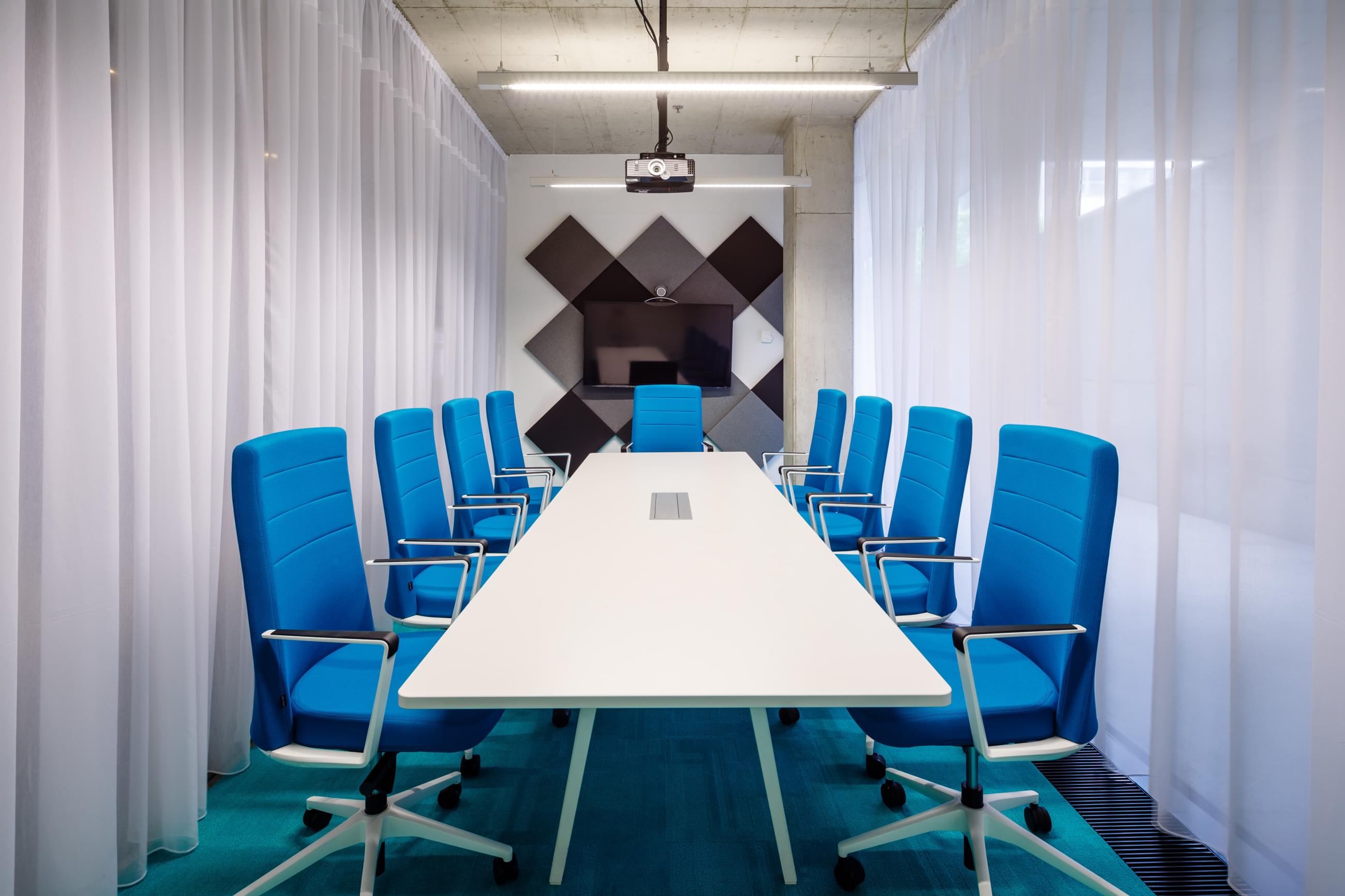 Zasedací místnost v modrých odstínech s bílým konferenčním stolem, černošedými akustickými panely a otevřenou prosklenou stěnou směrem do ulice.