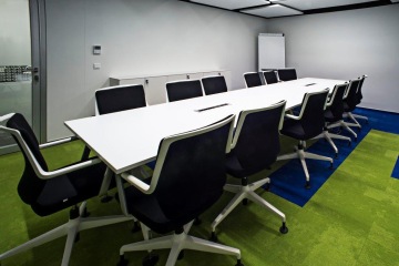 A meeting room where the dominant feature are green and purple carpet tiles and an elongated white meeting table with black and white office chairs.
