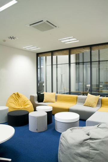 Pohled na relaxační zónu s šedo-žlutou pohovkou. Přes chodbu, která je z obou stran oddělená prosklenou stěnou, je zasedací místnost