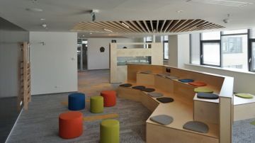 Vyvýšené pódium v relax zóně, vhodné např. pro neformální setkání, je vyrobeno z překližky a doplněno o barevné čalouněné podsedáky.