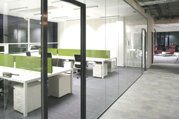 Kanceláře jsou oddělené bezrámovým sklem. Čalouněné akustické panely mezi stoly jsou zelené.