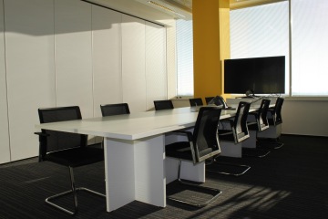 Meeting room with big LCD screen behind which is large window and inside the room is also dominant oblong white table in combination with black office chairs.