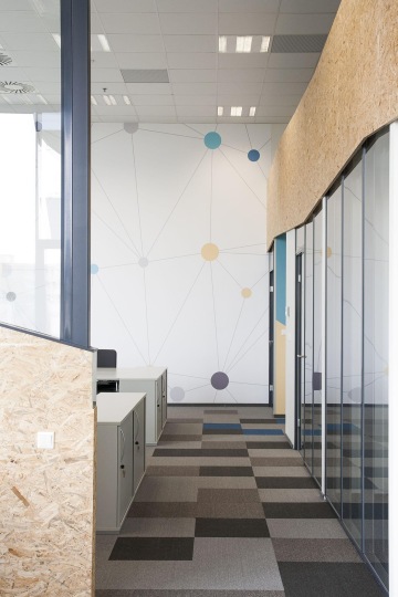 Pohled na velkoplošné tapety s barevnými motivy na konci chodby, kde na pravé straně jsou i prosklené stěny a po levici OSB deska s popisovatelným sklem.