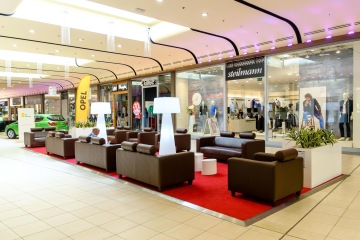 Kreativní relax zóna v OC Nisa Liberec je vybavena hnědými koženými křesly a bílými lampami, vše doplňuje kontrastní červený koberec a rostliny v bílých květináčích.