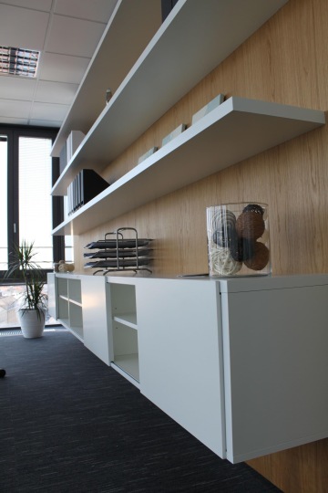 Bílý policový systém na dřevěné stěně v DELL showroomu. Prostoru dodává světlo velká prosklená stěna.