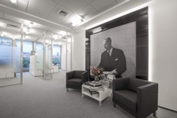 The new office at the Charles Square Center has large windows overlooking the city center, with gray modern armchairs and a white coffee table. The dominant black and white portal with the founder of Christian Dior stands out in the background.