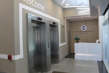 Light reception with white counter and logo on the wall, elevators and EUROPEUM entrance rooms. On the ceiling are modern narrow lights.