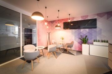 Šedá křesílka s malými stolečky jako místo k setkávání s LCD obrazovkami a zajímavě řešeným osvětlením a barevnou tapetou v pozadí kontrastní k jinak šedému interiéru.Šedá křesílka s malými stolečky doplňuje zajímavě řešené osvětlení a barevná tapeta, která je kontrastní k jinak šedému interiéru.