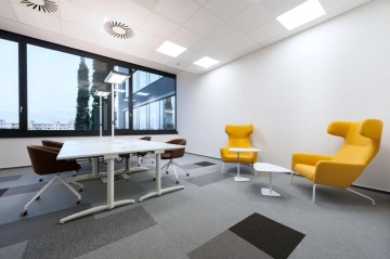 Mobilní stoly TRAMA se sklopnou deskou, doplněné o hnědá kancelářská křesla a kousek opodál o moderní křesla se zaoblenými hanami, která jsou určena k relaxaci.