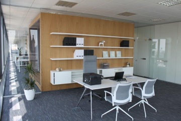 Bílý praktický stůl a šedé židle z kolekce nábytek ACTIU v moderním DELL showroomu. V pozadí je dřevěná stěna se skleněným detailem.