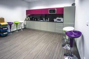 The kitchen has gray lower cabinets and purple upper cabinets, there is white, green and purple bar chairs too.