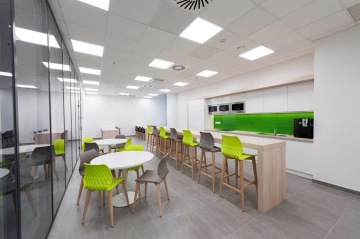 Prostorná kuchyňka s velkým počtem zelených a šedých židlí v kombinaci klasických a barových s přilehlými stoly a stropním osvětlením.