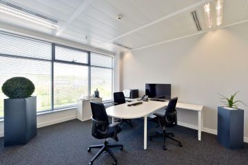 Large windows go through Regus' office, a black upholstered office chair with a metal construction around the round white desk. The room is complemented by a green plant.