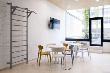 Pohled na část místnosti určené k relaxaci, vybavené kulatými stolečky s barevnými židlemi, LCD televizí na stěně a žebřinami k protažení svalů