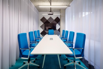 Meeting room in blue shades with a white conference table, black-colored acoustic panels and an open glass wall facing the street.