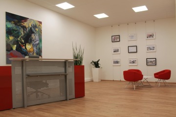 Showroom City Business Residence se vyznačuje několika druhy materiálů - dřevěná pracovní deska na recepci, červené čalouněné židle, bílý stolek s kovovou konstrukcí.