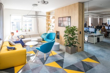 Pohled na relax zónu ve společnosti CAPEXUS. Pohodlné pohovky ve světlých barvách s tyrkysovými křesílky a bílými stolečky.