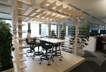 Neobvyklý design open space kanceláře skvěle doplňují odstíny světlých barev. V kancelářském prostoru se také nachází dřevěná konstrukce vyrobená ze světlého dřeva. V popředí je pracující zaměstnanec firmy.