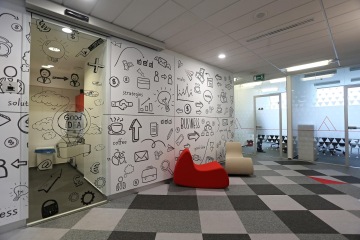 The space in front of the kitchen is complete with beige and red upholstered chairs. On the wall is a large-format wallpaper with pictograms and on the floor is gray carpet squares alternating with red carpet triangles.