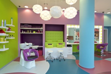 Funny Sussy baby hairdresser is pleasantly enlightened by round white lamps. The interior features purple chairs and shelves and a green sofa.