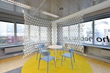 V prostoru Showroomu Zlatý Anděl se nachází modré jednoduché židle a bílý stolek navržení společností ACTIU, žlutý koberec a bílá designová clona dodávají místnosti příjemnou atmosféru.