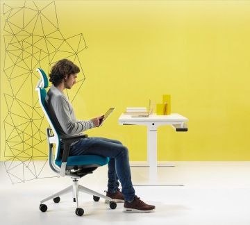 Minimalistická místnost se žlutou stěnou v popředí s mužem pracujícím na tabletu. Muž sedí na tyrkysové kancelářské židli TNK-Flex, která splňuje všechny ergonomické zásady.
