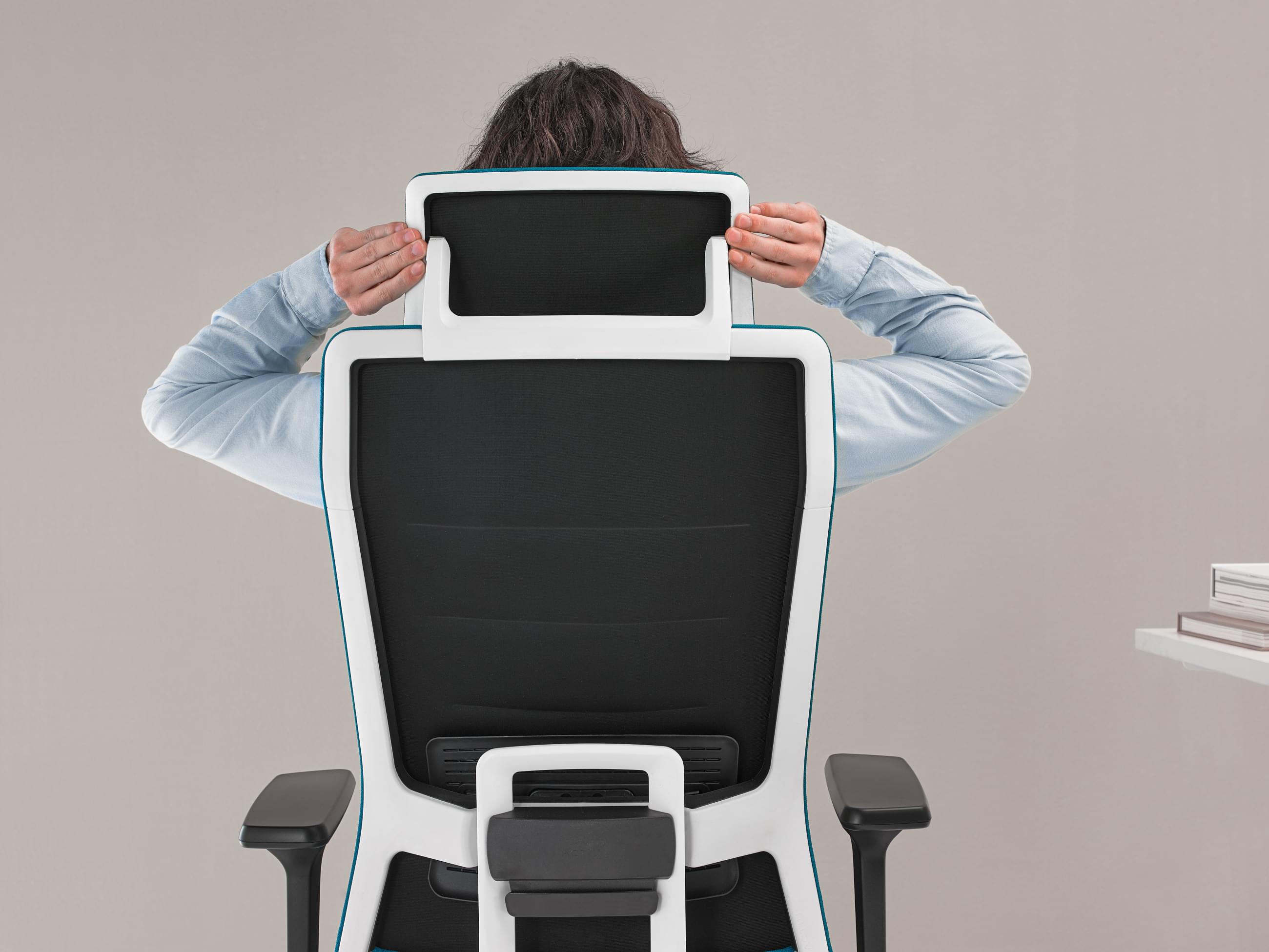 Kancelářská židle s tyrkysovou barvou má ergonomické prvky pro zdravé záda. Na obrázku lze vidět zaměstnance nastavující opěrku hlavy. 